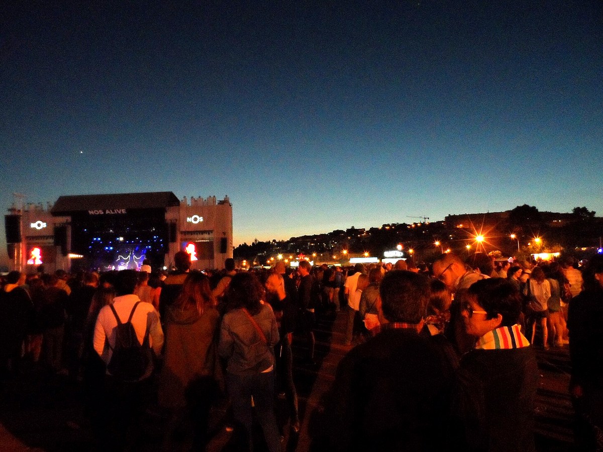Público de um festival ao anoitecer, com o palco iluminado ao fundo